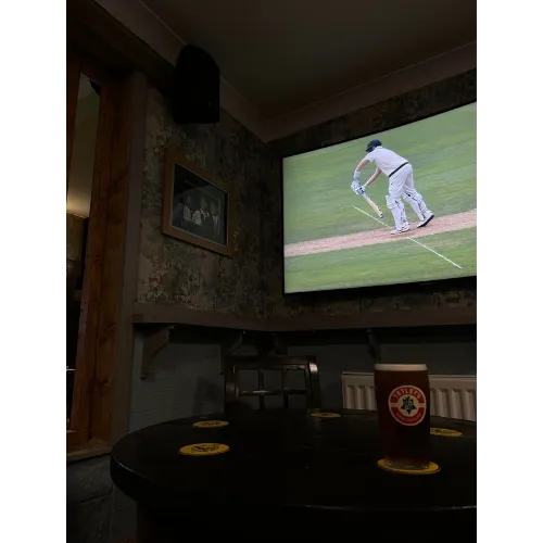 A television in a pub displays a cricket match while a man enjoys a beer nearby, creating a lively atmosphere.